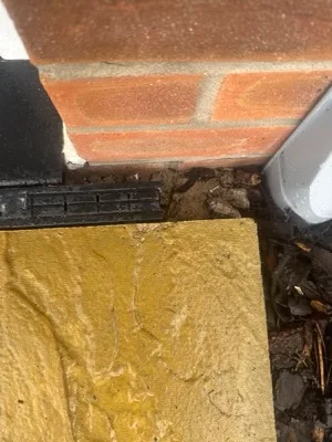 Bare earth between a buff paving slab and the brick door reveal, where the path has not been finished.